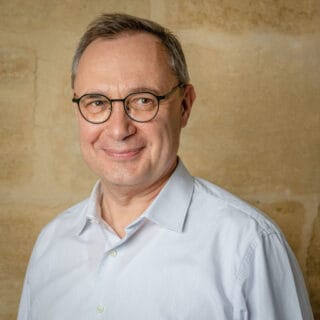 Jean-Philippe JOUBERT, a middle-aged man with short gray hair and glasses, is smiling warmly. He is wearing a light blue button-up shirt and standing in front of a tan, textured wall.