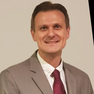 A man with short brown hair, wearing a gray suit, white shirt, and red tie, is smiling against a neutral background.