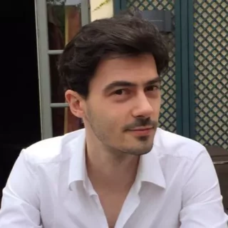 A man with short dark hair and a trimmed beard is sitting outdoors, wearing a white button-up shirt. Behind him, there is a lattice fence, and part of a building door is visible. He has a slight smile and is looking towards the camera, possibly contemplating his next move in the SNITEM project.